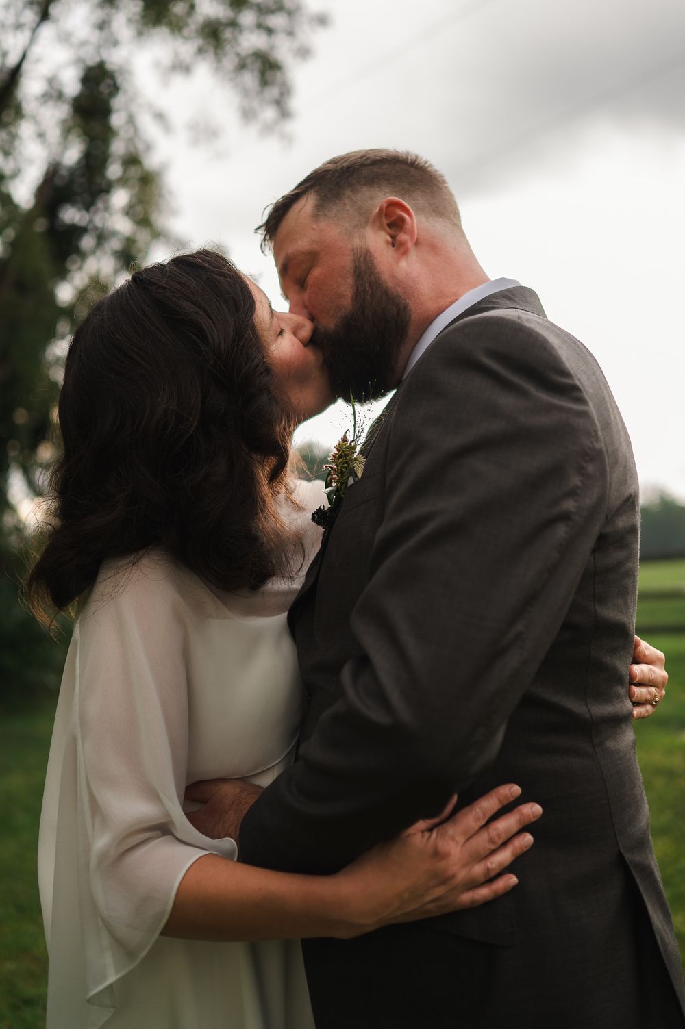 A bride and groom share a romantic kiss outdoors on their wedding day, surrounded by lush green trees under a cloudy sky.