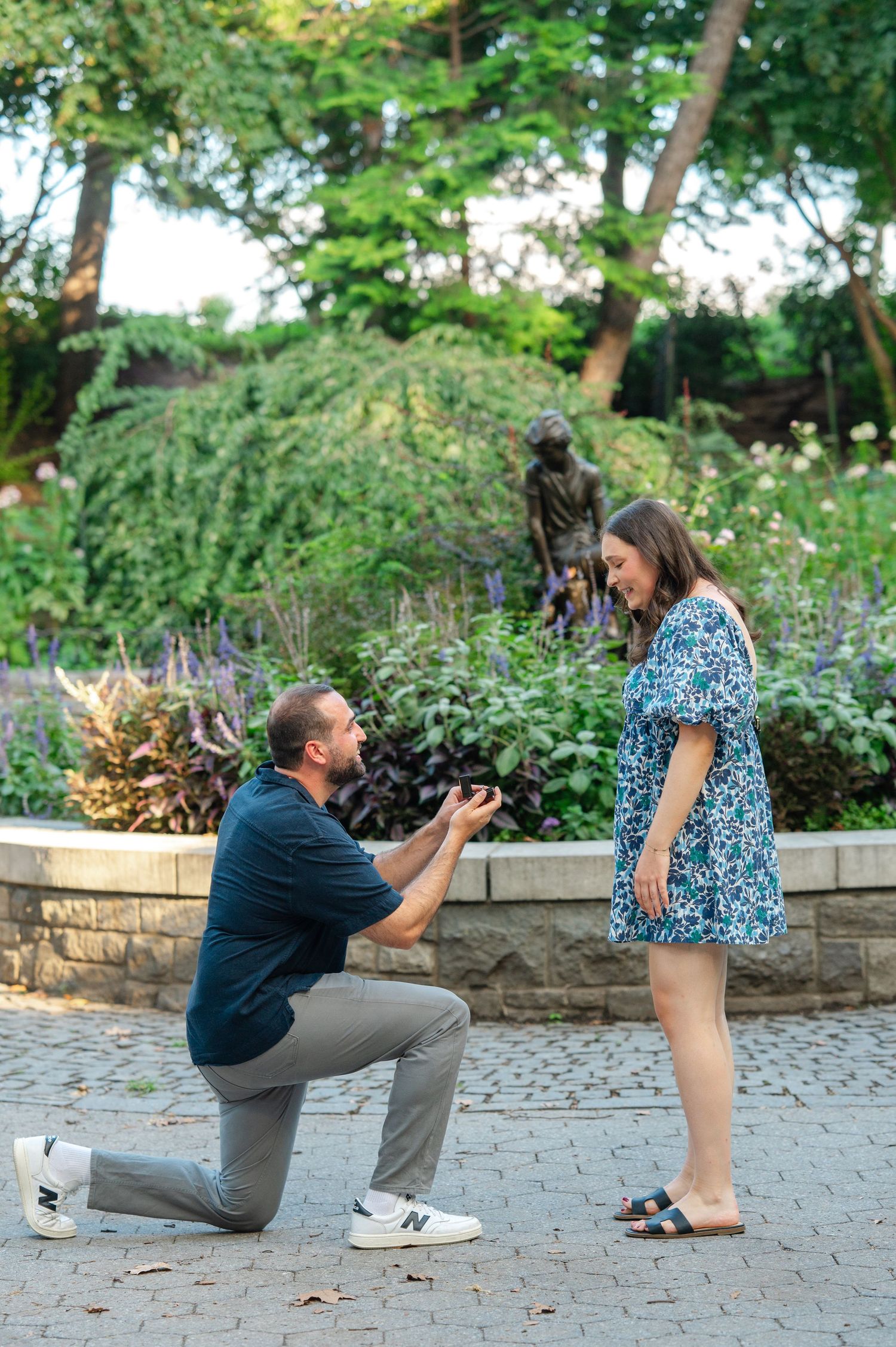 Man proposes on one knee to woman in floral dress in a lush garden park setting of Carl Schurz Park on the UES with statue backdrop.