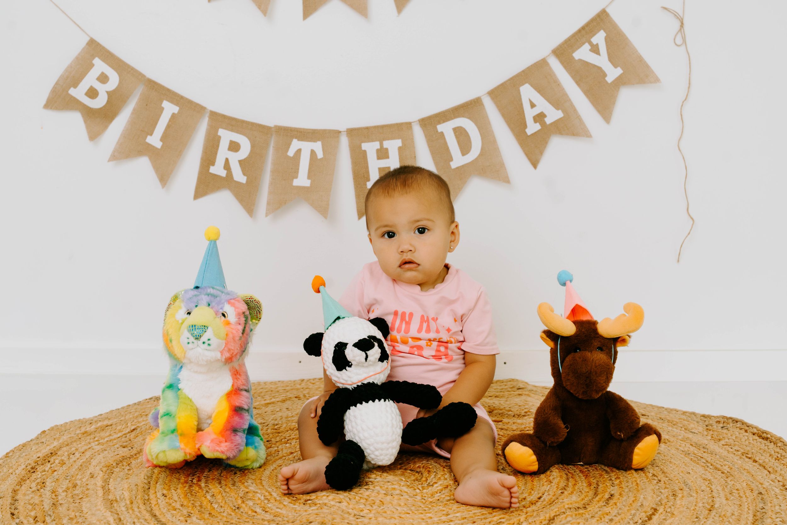 Baby in pink shirt sits between colorful stuffed animals with party hats under a Birthday banner.