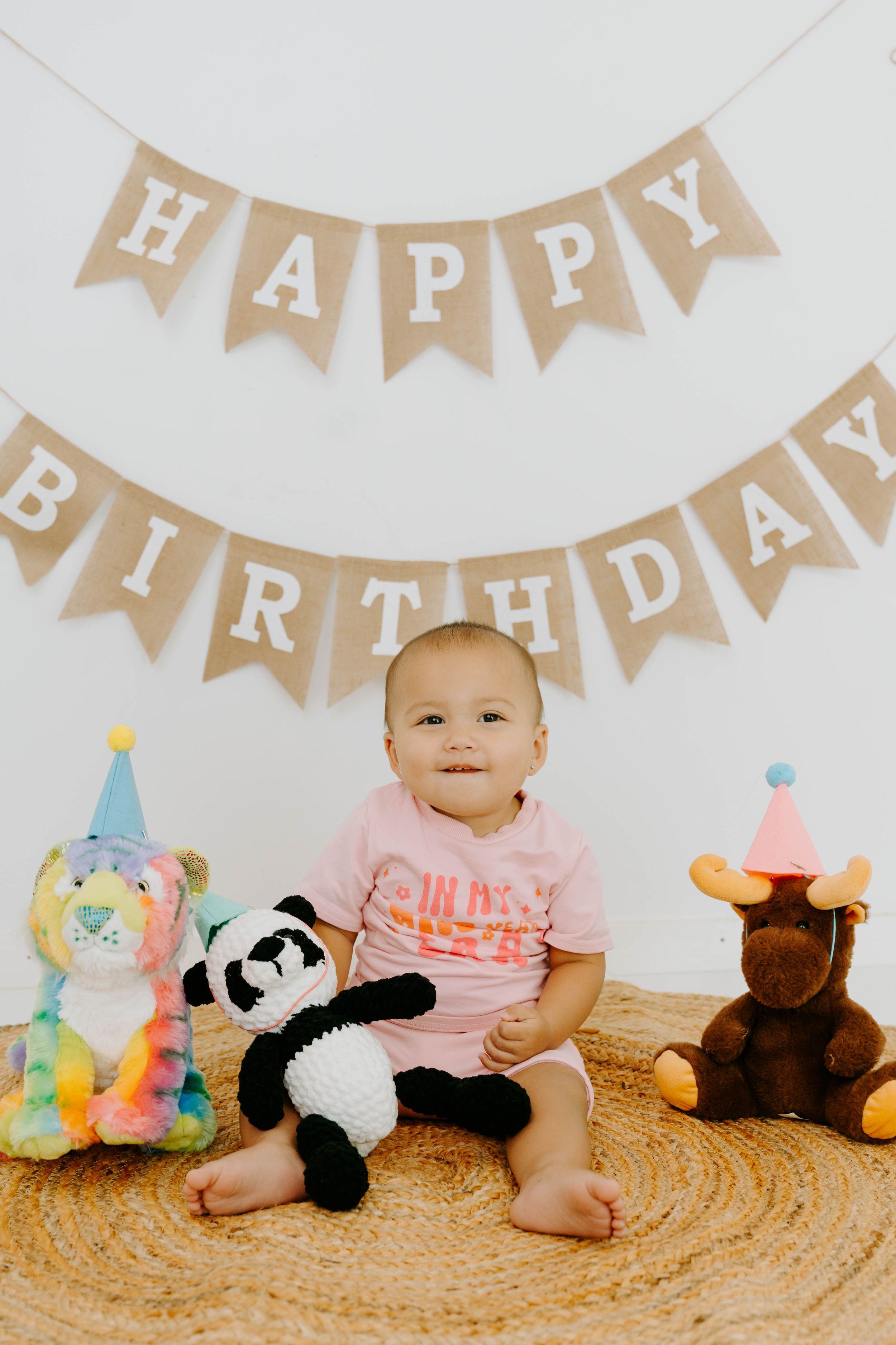 Cute baby in pink shirt sits with stuffed animals under a Happy Birthday burlap banner.