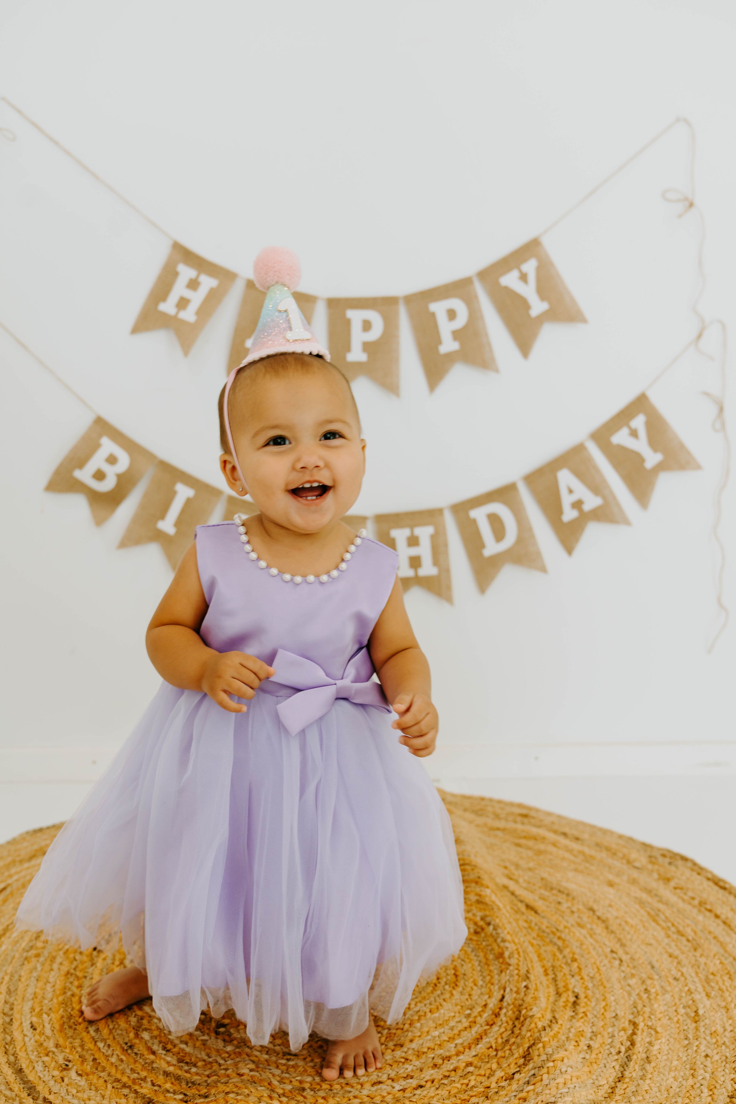 A smiling baby in a lavender tutu dress and birthday hat stands before a Happy Birthday burlap banner.
