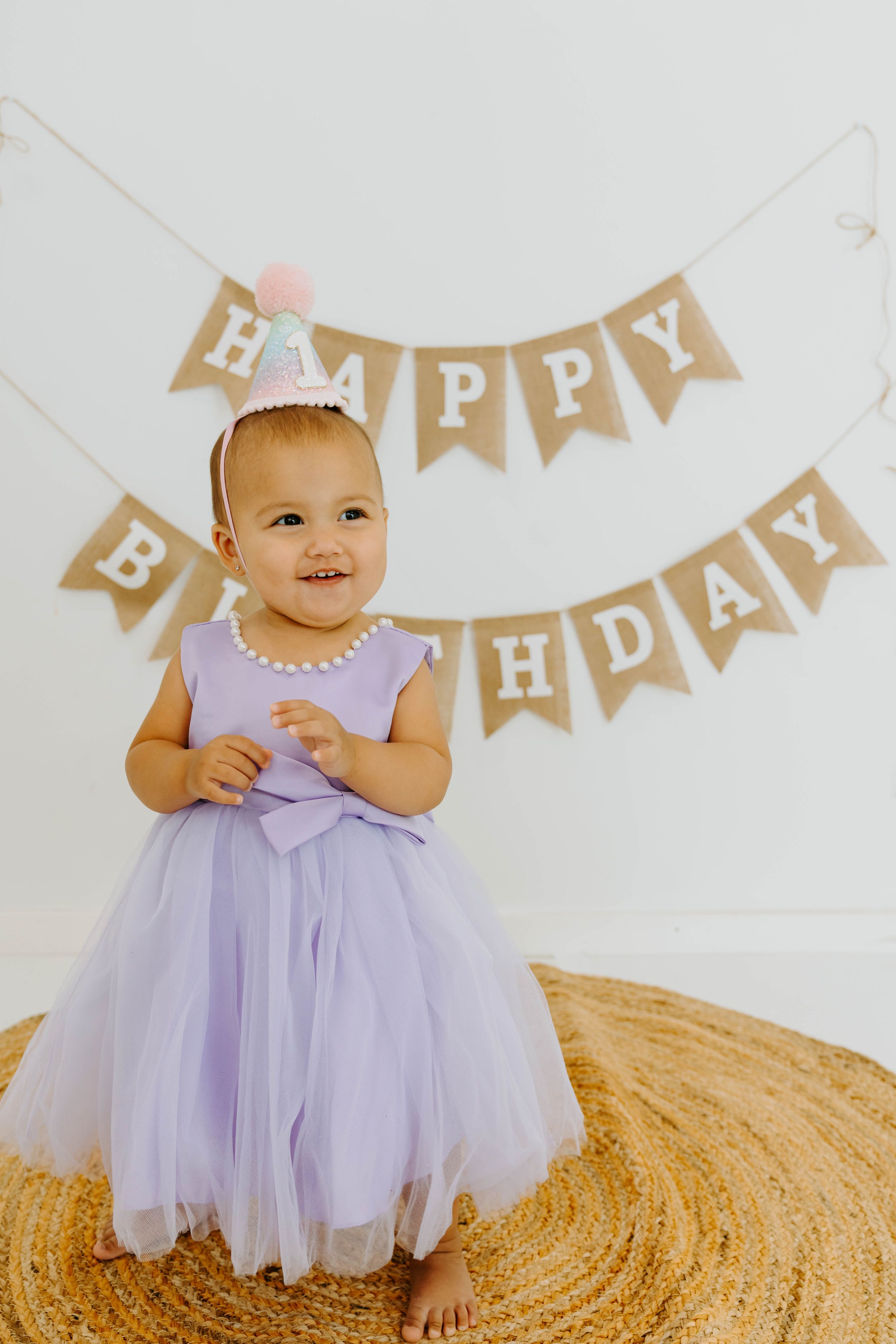 Smiling baby girl in lavender tutu dress and birthday crown sits before Happy Birthday banner.