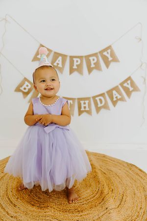 Smiling baby girl in purple tutu dress and party hat stands before Happy Birthday banner on woven rug.