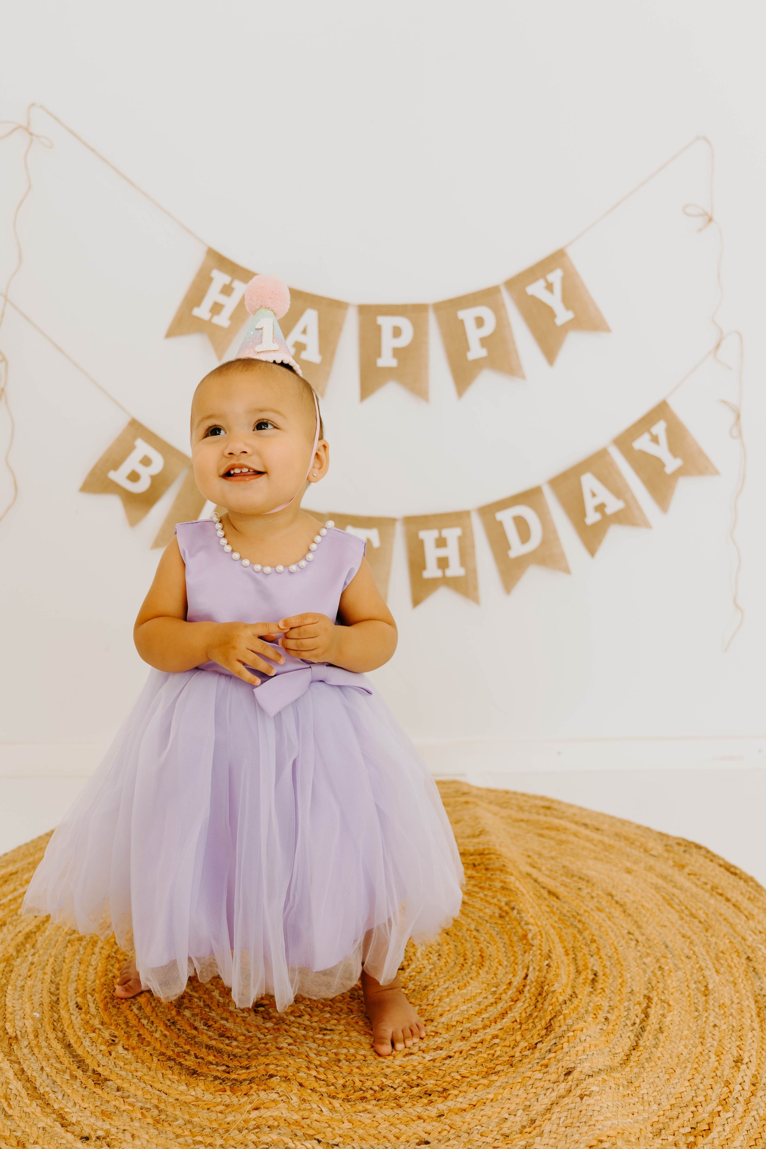 Adorable toddler in purple tulle dress and party hat stands before burlap Happy Birthday banner.