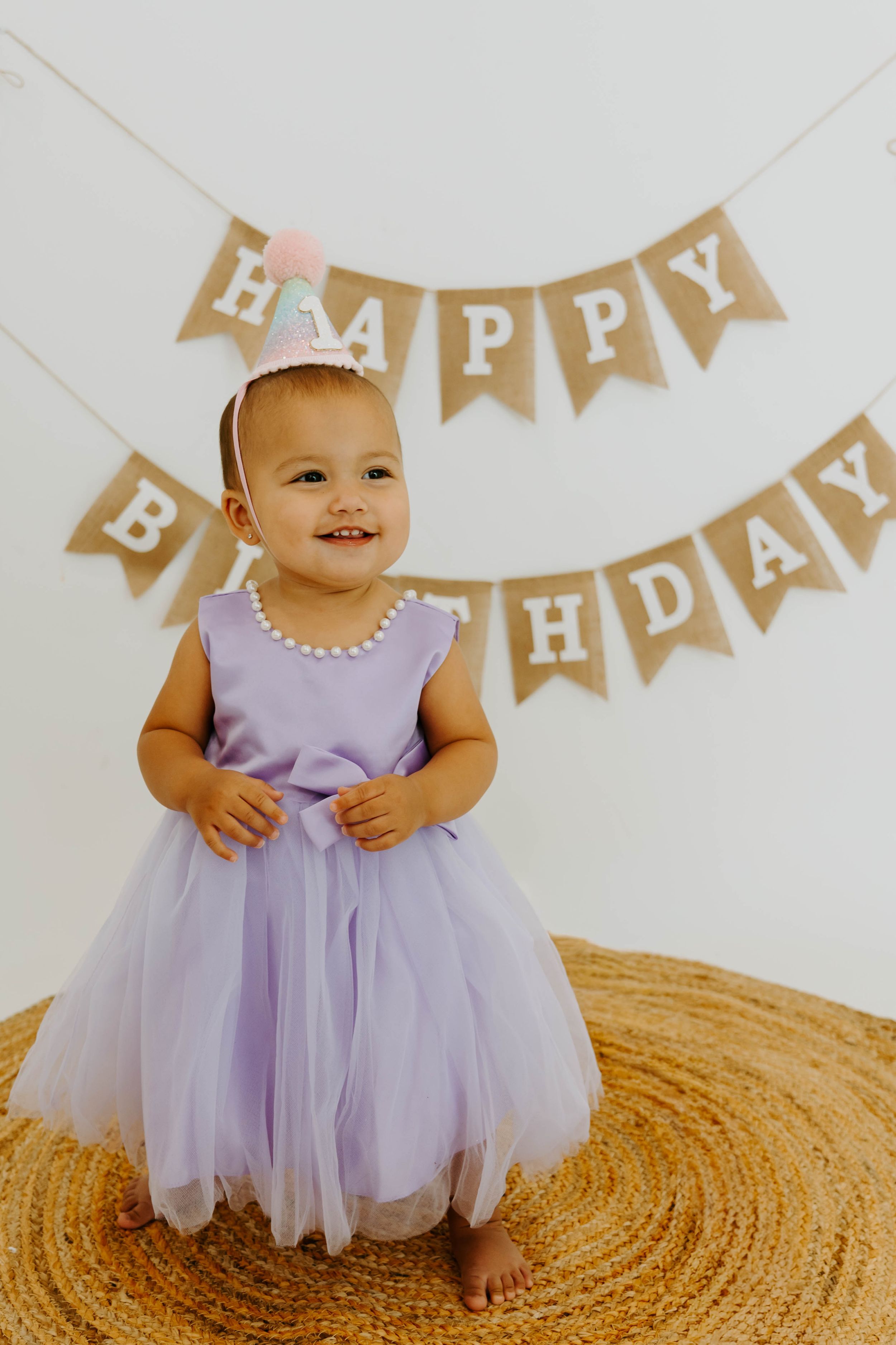 Smiling baby girl in purple tulle dress and birthday crown stands before a Happy Birthday burlap banner.