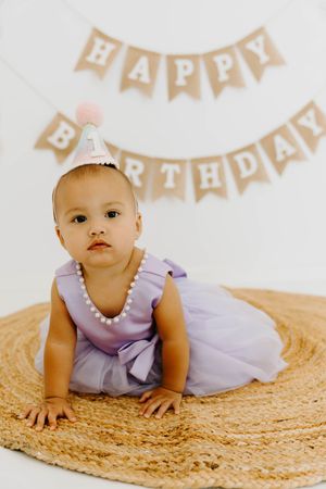 Baby girl in lavender dress and party hat crawls on woven rug in front of Happy Birthday banner.