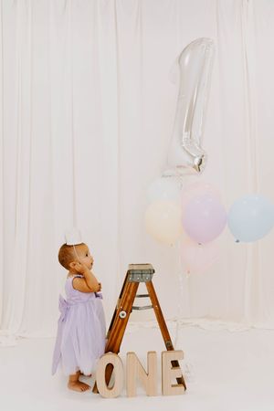 Toddler in lavender dress stands by wooden ladder with ONE letters and number 1 balloon at first birthday.