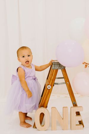 Baby girl in lavender dress stands by wooden ONE letters and balloons at first birthday photo shoot.