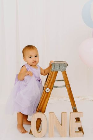 Baby girl in lavender dress stands by wooden ladder with ONE letters for first birthday photo session.
