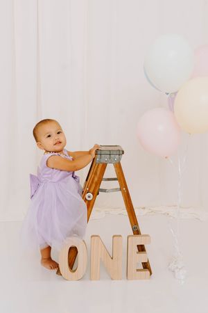 Baby girl in purple tutu dress stands by wooden ladder with ONE letters and pastel balloons for first birthday.