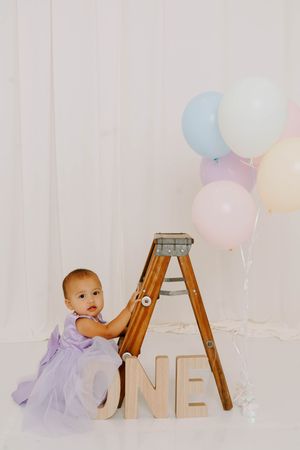 Baby girl in lavender dress sits by wooden ladder with ONE letters and pastel balloons for first birthday photo.