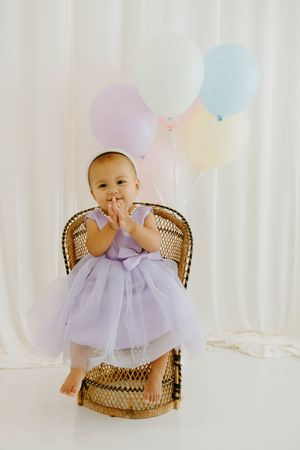 Baby girl in lavender tutu dress sits in wicker chair surrounded by pastel balloons.