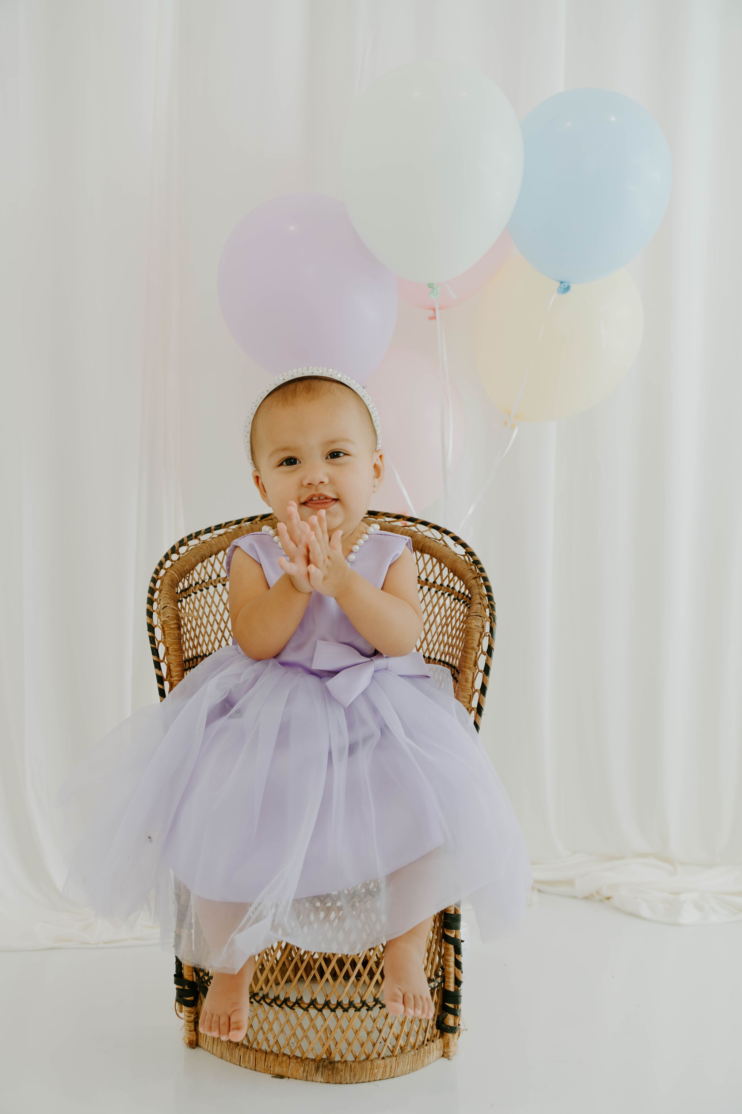 Baby girl in lavender tutu dress sitting in wicker chair with pastel balloons, clapping hands and smiling.