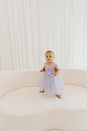 Cute baby girl in lavender tutu dress stands on white curved sofa against white curtain backdrop.
