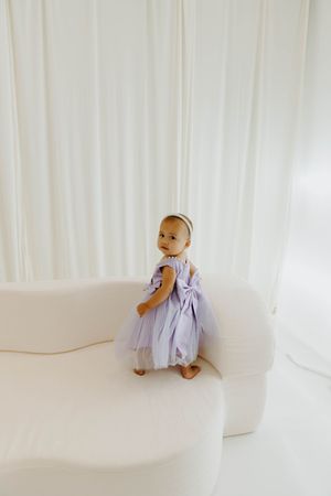 Baby girl in lavender tulle dress with bow stands on cream sofa against white curtain backdrop.