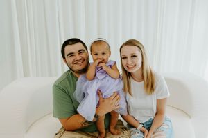 Happy young family with parents smiling and holding their baby girl in a purple dress on a white couch.