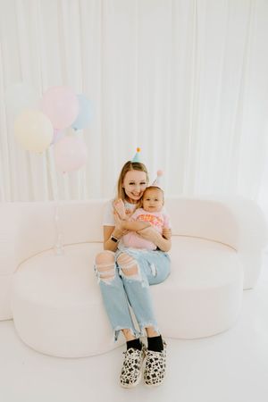 A young woman sits on a white sofa holding a baby in a party hat, surrounded by pastel balloons.