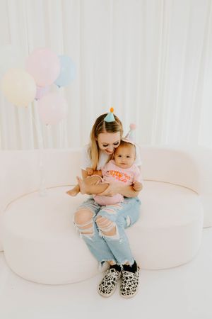 Mother in ripped jeans holds baby in pink outfit on white couch surrounded by pastel balloons.