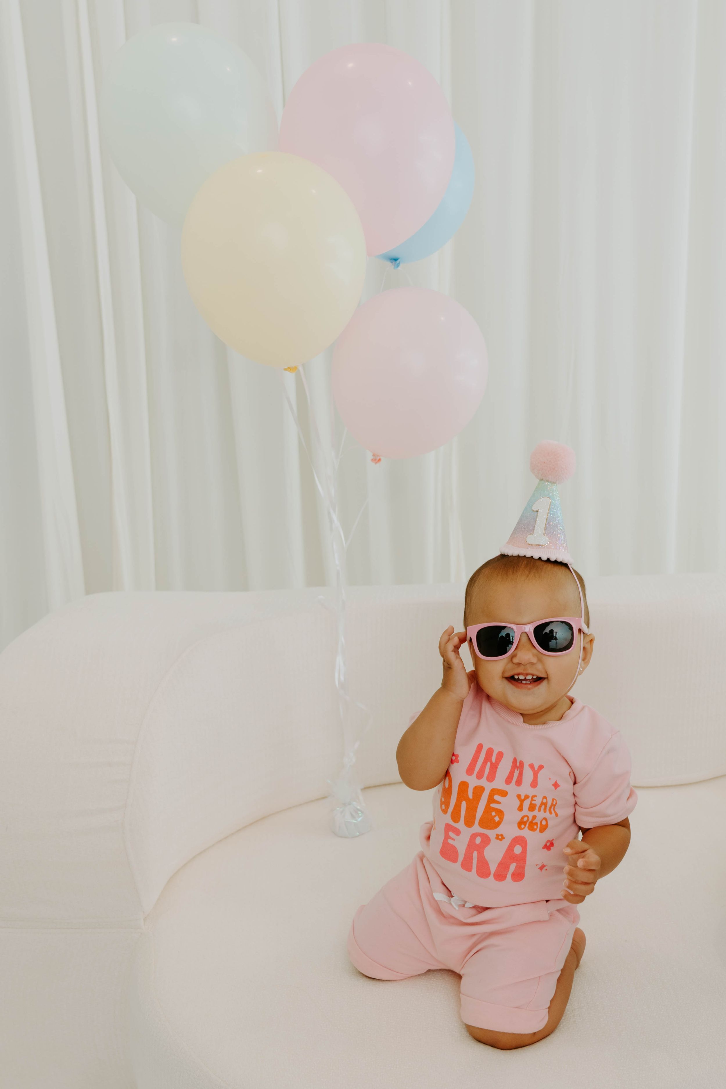 Baby girl in pink outfit, sunglasses, and party hat sits with pastel balloons celebrating her first birthday.