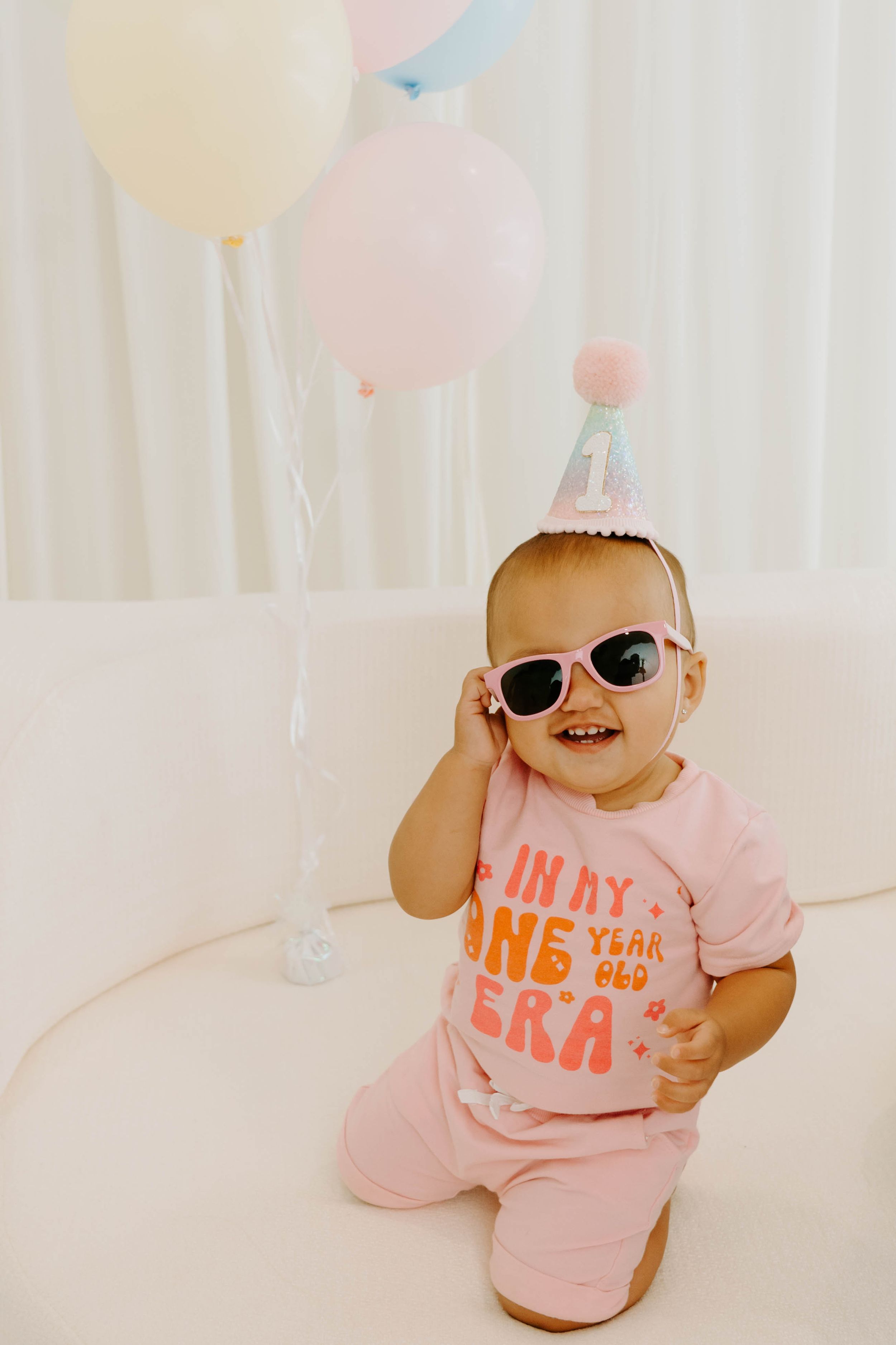 Baby in pink \"One Year Old Era\" shirt, sunglasses, and party hat poses with pastel balloons.
