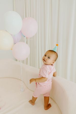 Adorable baby in pink outfit and party hat standing by pastel balloons on a white sofa.