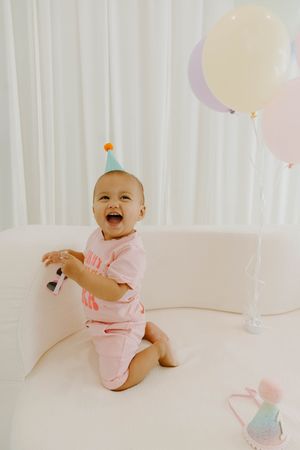 Laughing baby in pink outfit and party hat sits on white sofa with pastel balloons nearby.