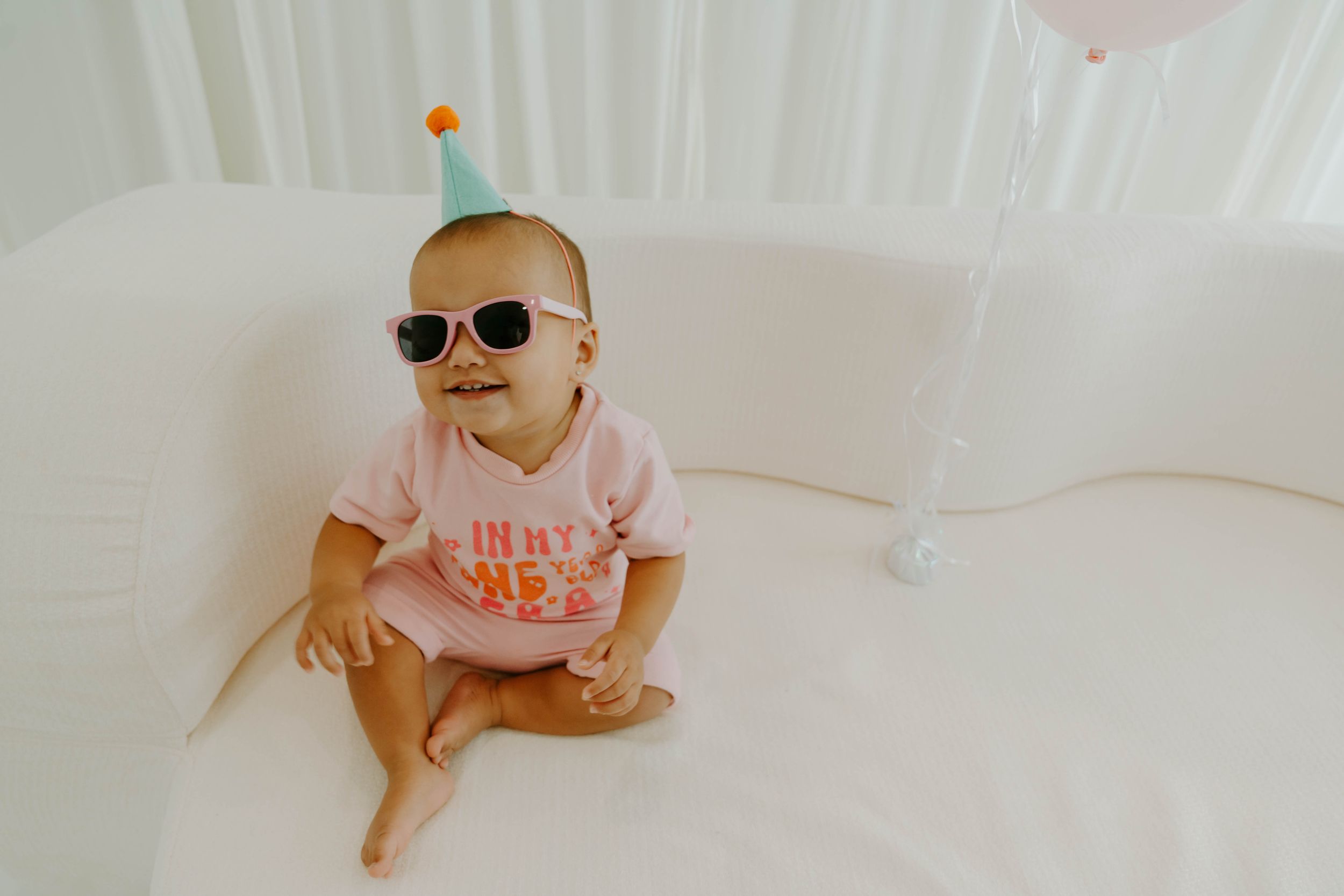 Cute baby wearing sunglasses and a party hat sits in a white crib celebrating their first birthday.