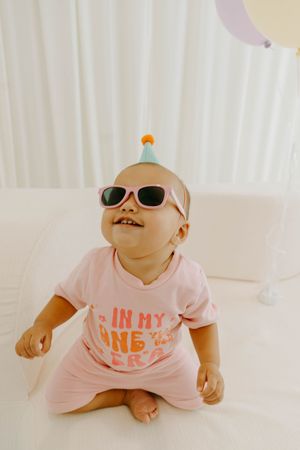 Adorable baby wearing pink sunglasses and birthday hat, dressed in pink outfit, sitting on white surface.