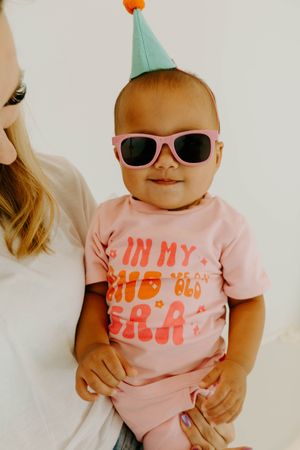 Baby wearing pink sunglasses and party hat held by mom in pink shirt at birthday celebration.