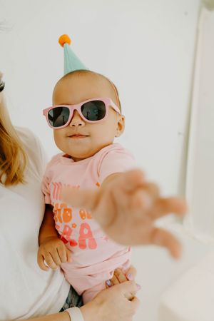 Baby wearing pink sunglasses and a party hat being held, reaching toward camera on first birthday.