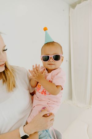 Adorable baby wearing pink sunglasses and a party hat being held by a woman in a white shirt.