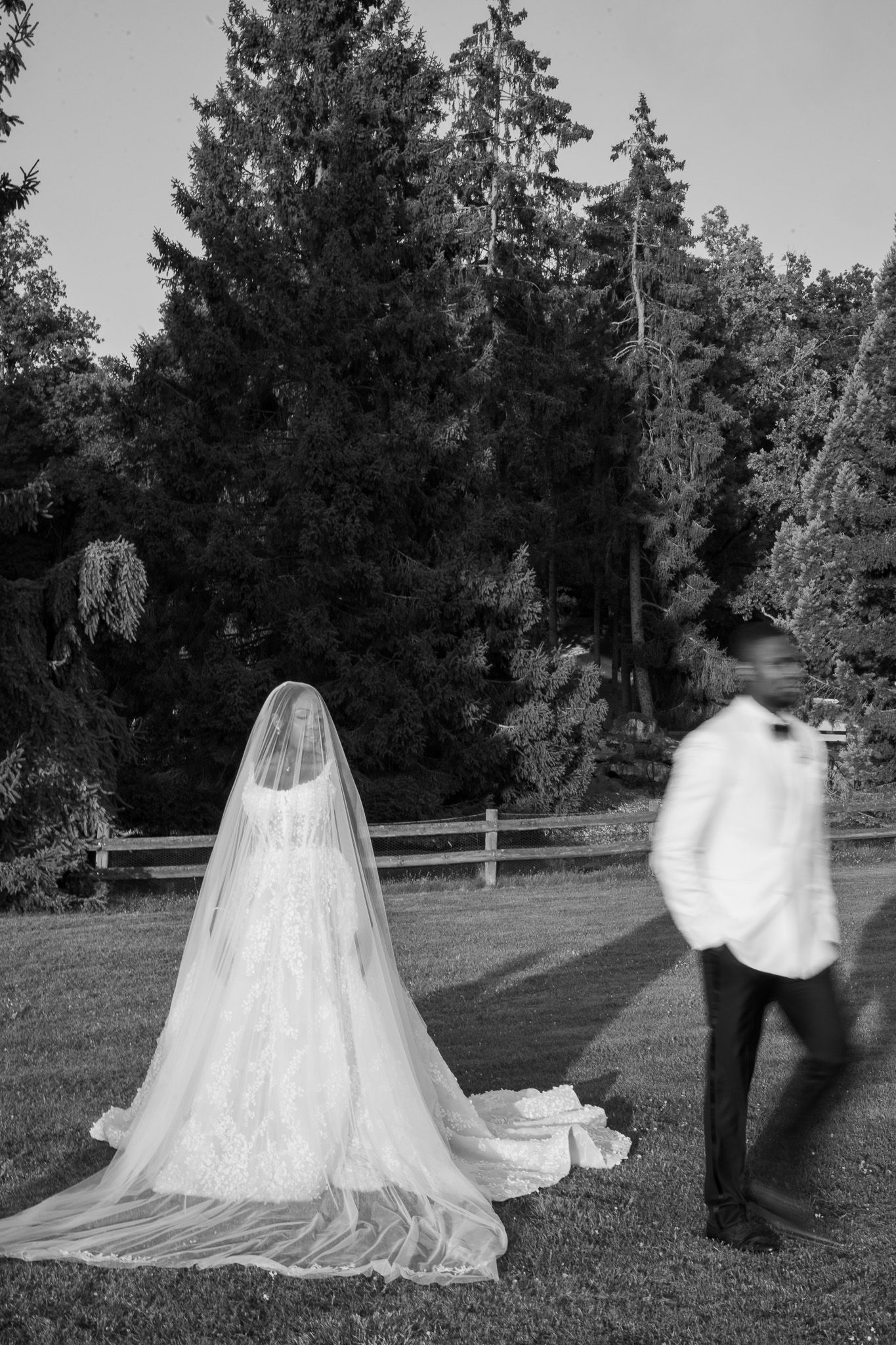 Black and white wedding photo of bride in flowing gown and veil with groom in white jacket outdoors.