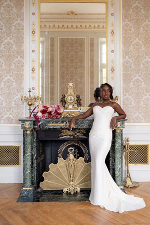 Elegant bride in white mermaid gown poses by ornate green marble fireplace in luxurious gold-adorned room.