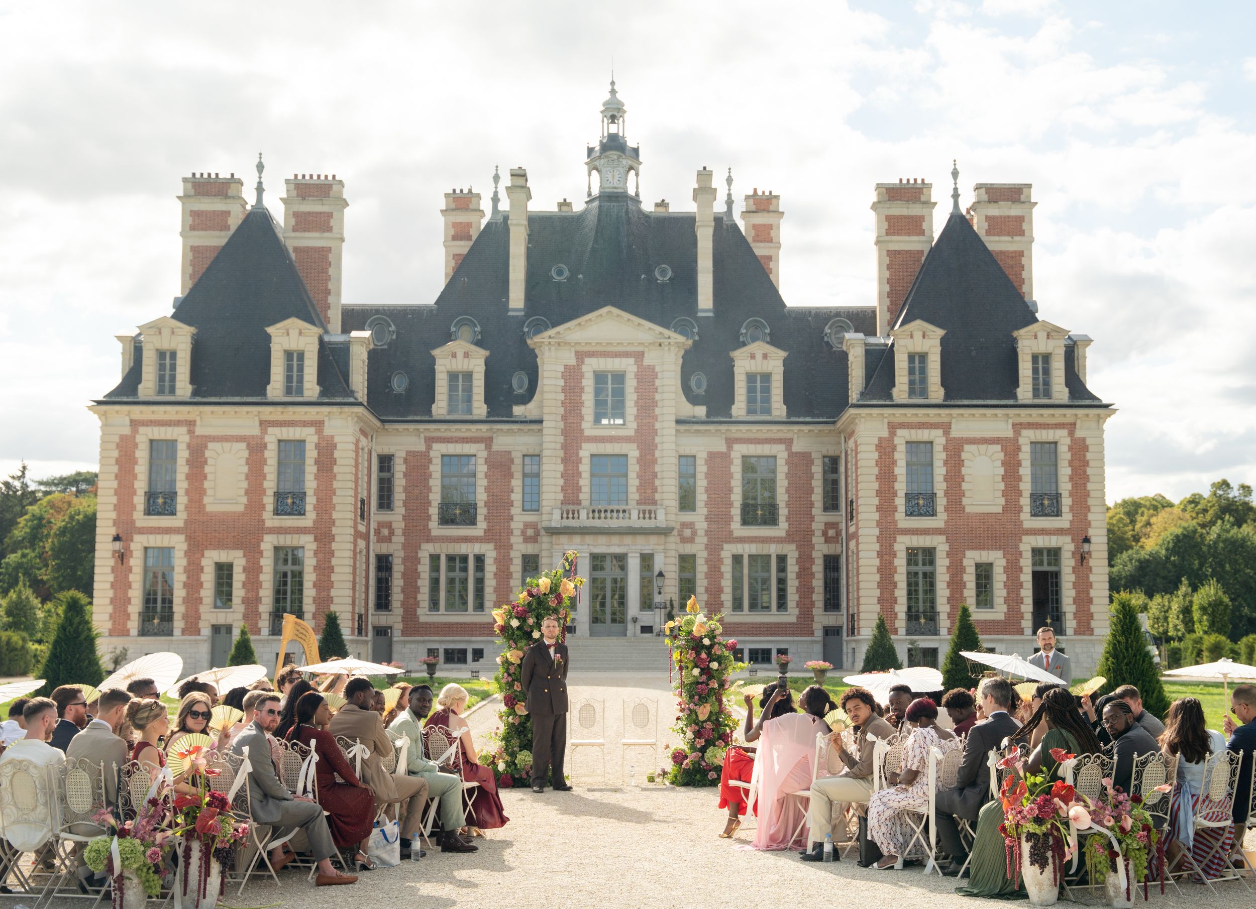 Elegant outdoor wedding ceremony at a grand French château with guests seated on a sunny terrace.