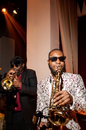 Two musicians perform jazz, one playing trumpet and another in a floral blazer playing saxophone under warm stage lights.