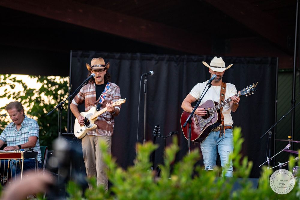 Two country musicians perform on outdoor stage, one playing electric guitar, another with acoustic guitar and cowboy hat.