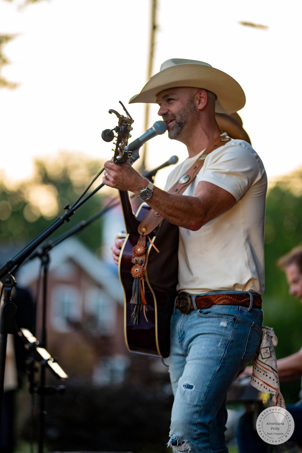 Country musician in cowboy hat plays acoustic guitar and sings into microphone on outdoor stage in jeans and white tee.