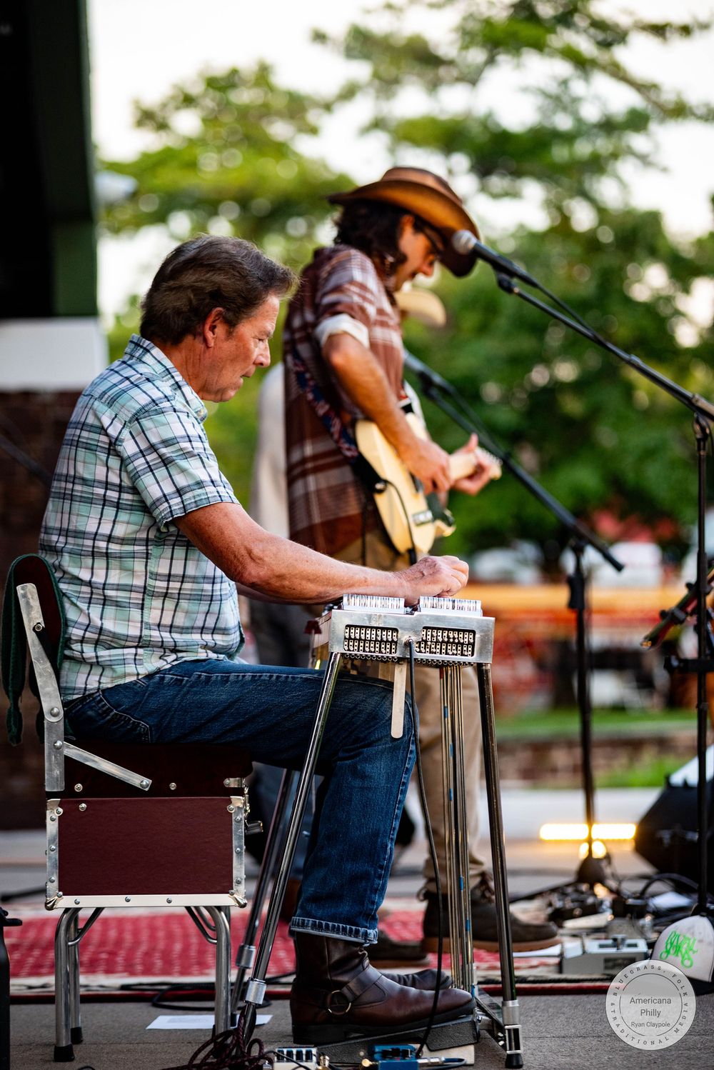 Musician playing pedal steel guitar seated on stage while guitarist performs in background outdoors.