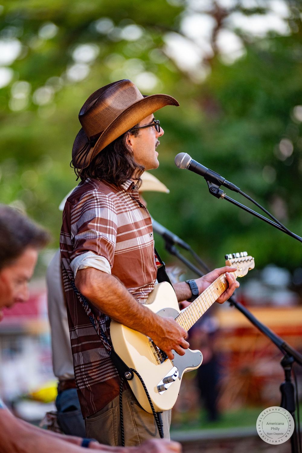 Musician in cowboy hat playing electric guitar and singing into microphone at outdoor concert.