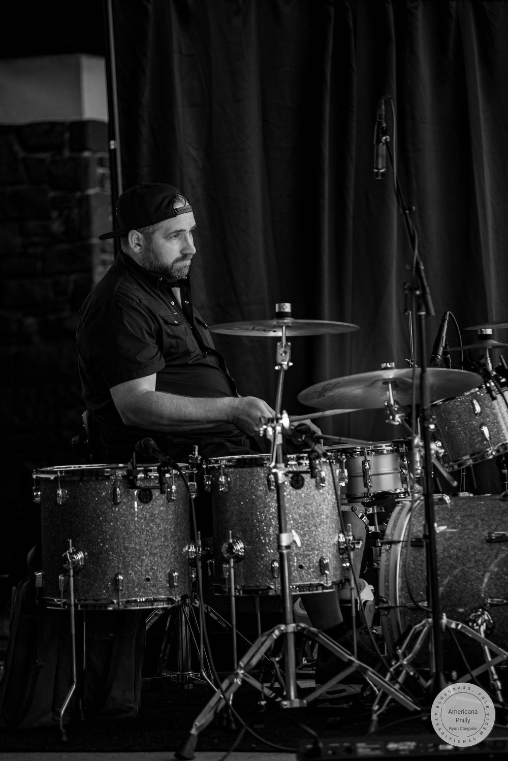 Black and white photo of a bearded male drummer performing on stage with a full drum kit.