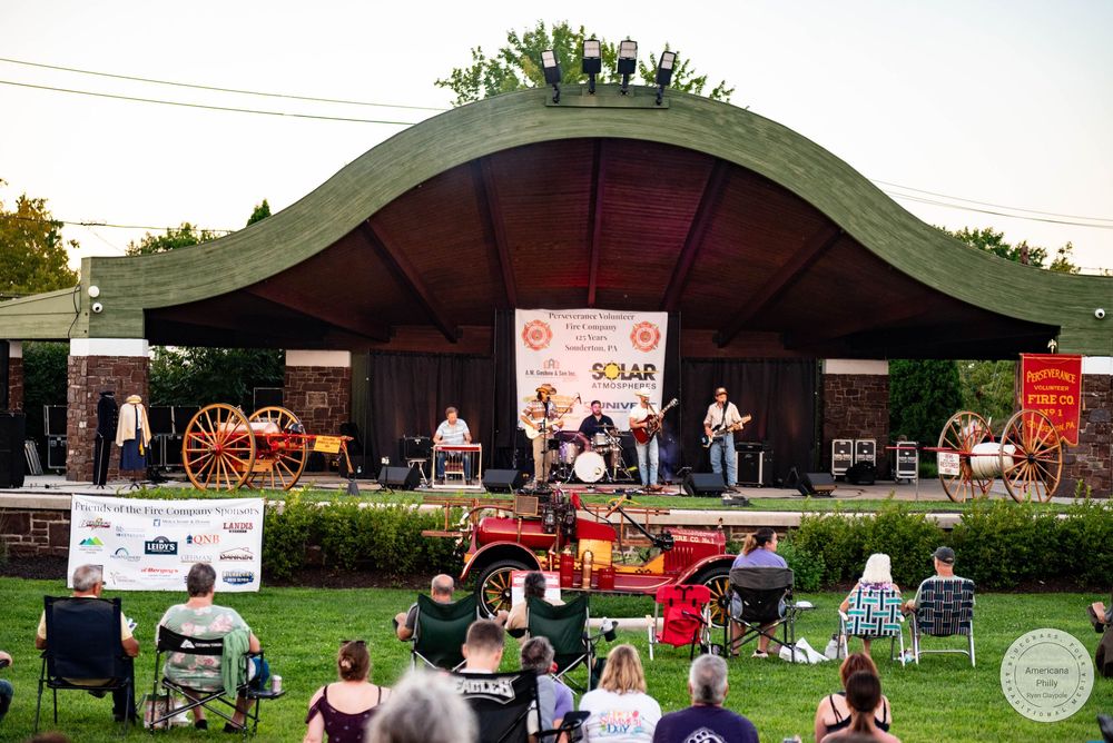 Outdoor country music concert at a park bandshell with audience seated on lawn chairs and grass.