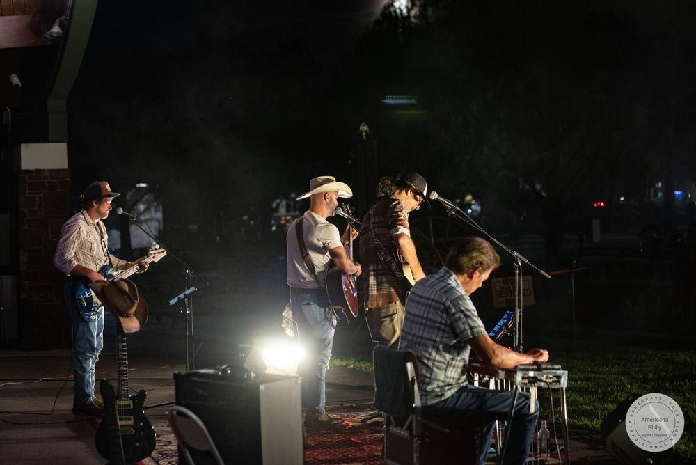 Country band performing outdoor night concert, musicians playing guitars and keyboard on stage with bright lights.