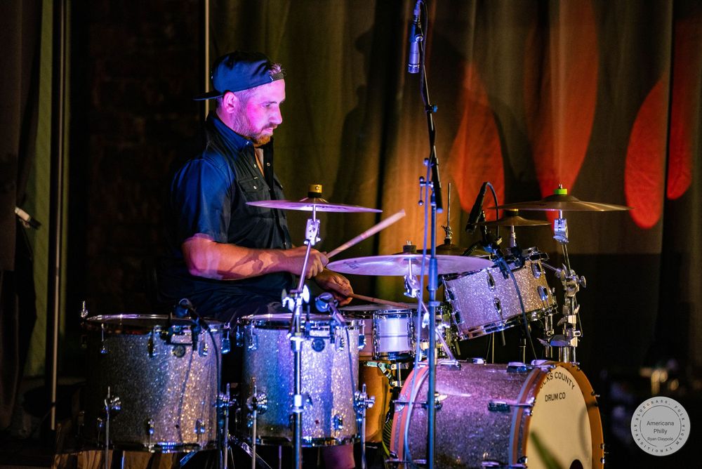 A male drummer performs on stage under colorful lights with a full drum kit setup during a live music event.