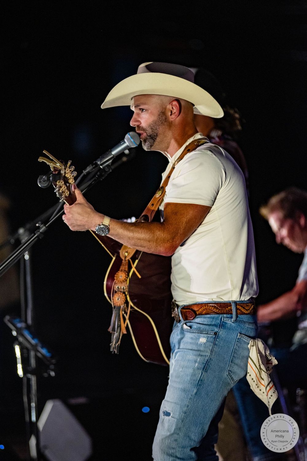 Country musician performing on stage with guitar and cowboy hat singing into microphone at outdoor concert.