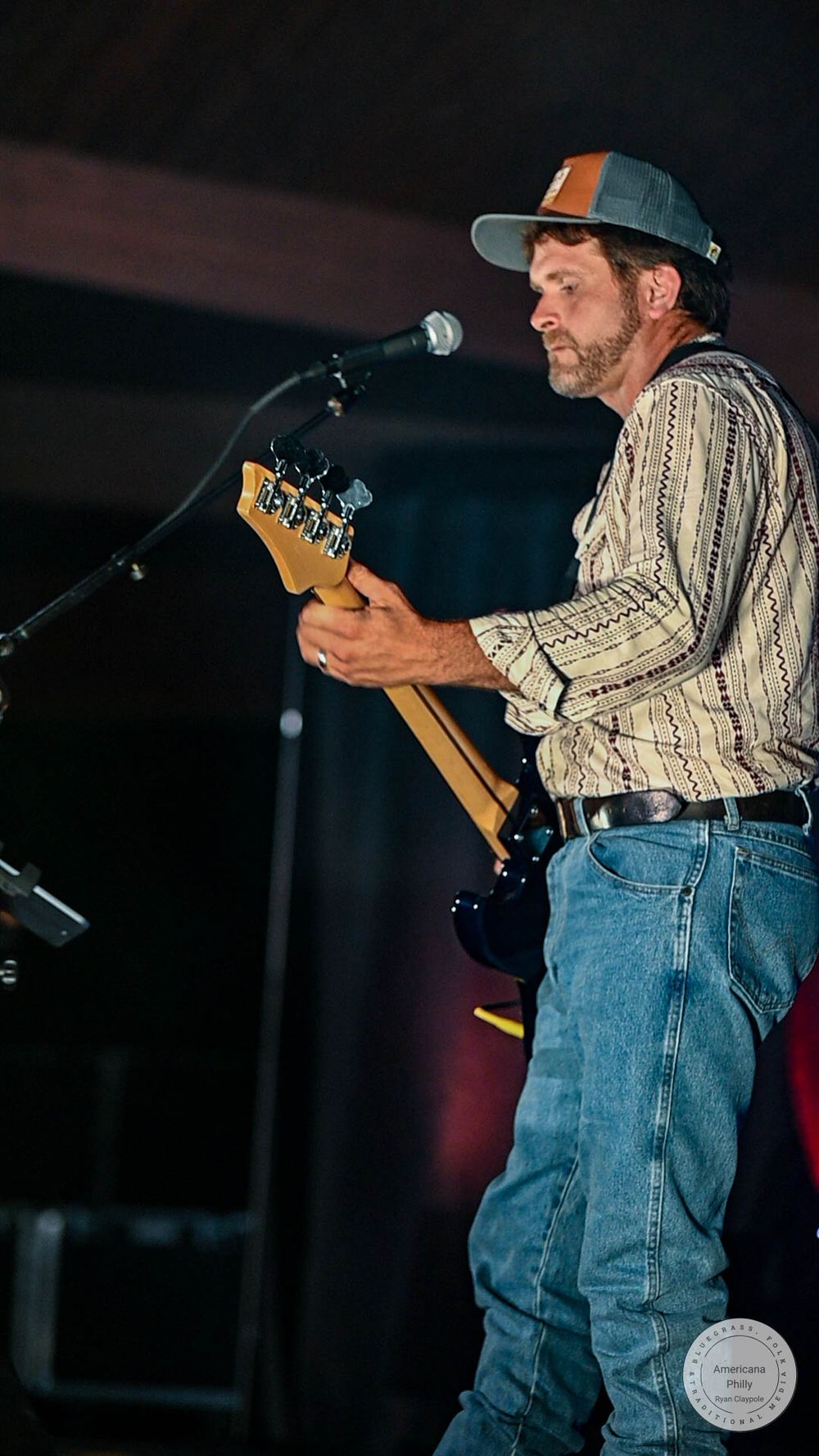 A male guitarist in a striped shirt and gray cap performs on stage with a microphone stand under dark lighting.