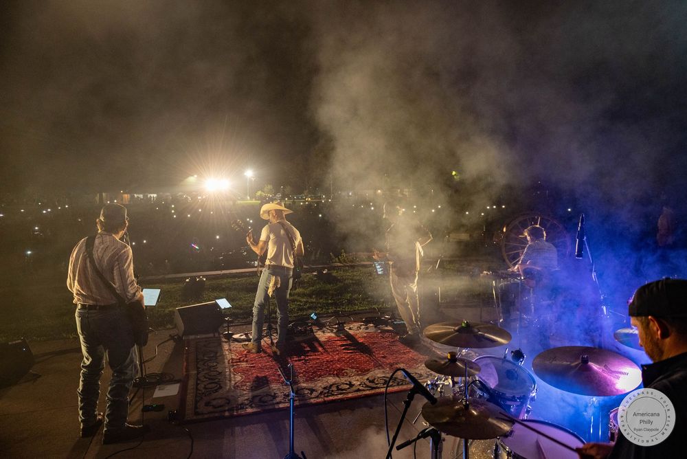 Country band performing on outdoor stage at night with dramatic fog and city lights in background.