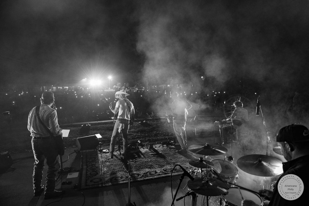 Black and white photo of a live band performing on an outdoor stage at night with dramatic smoke and crowd lights.