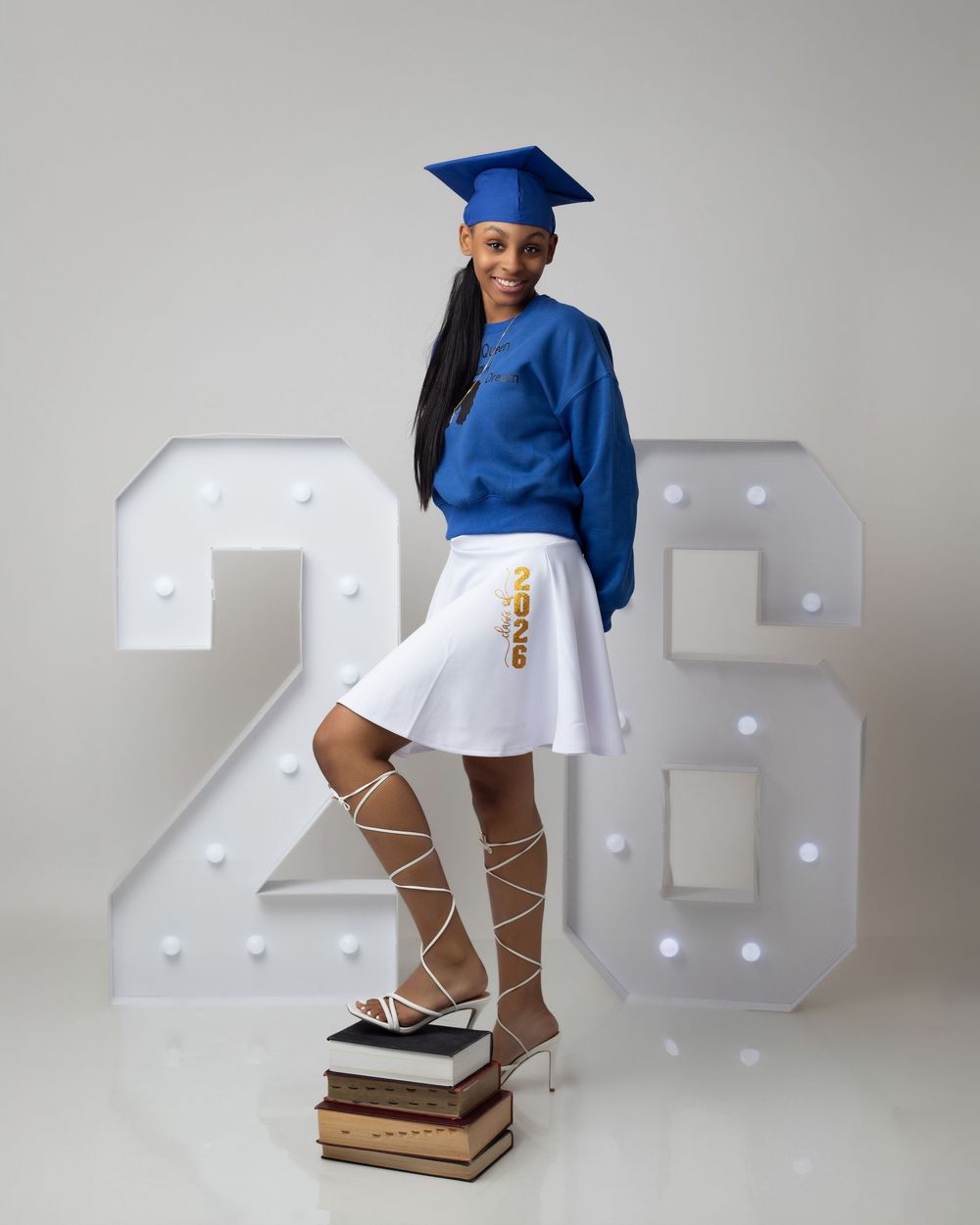 Graduate in blue cap and gown poses with books and illuminated '26' marquee letters for senior portrait.