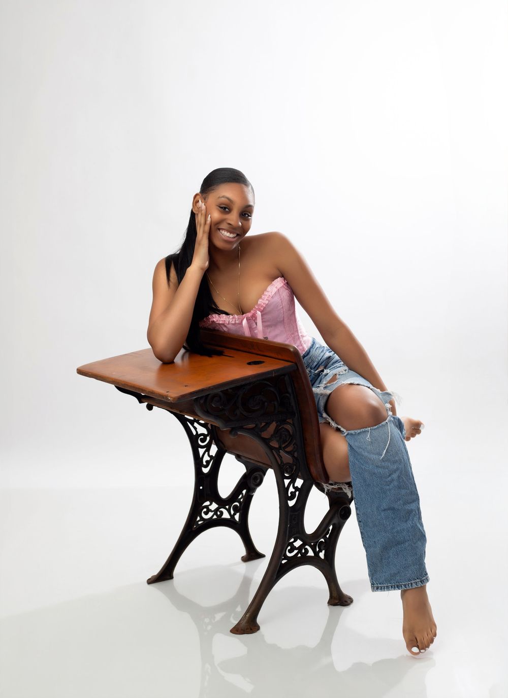 Young woman in pink top and ripped jeans sitting relaxed on a vintage school desk, smiling against white background.