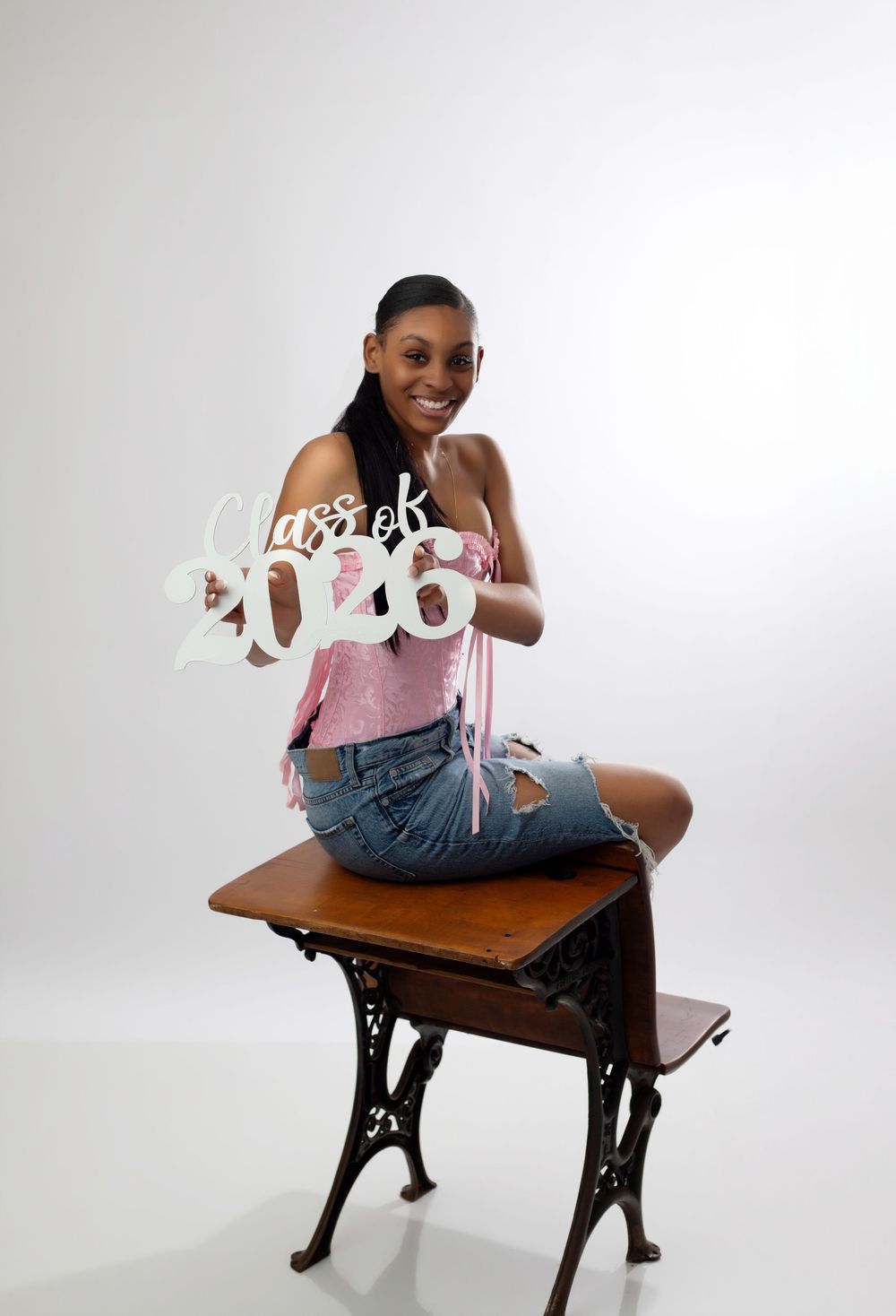 Young woman sitting on vintage desk holding Class of 2026 sign, wearing pink top and denim shorts.